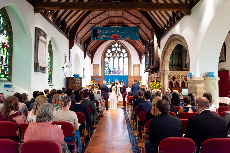 Ashley and Anjana look at eachother  surrounded by wedding guests during their wedding in Southover Church, Lewes.