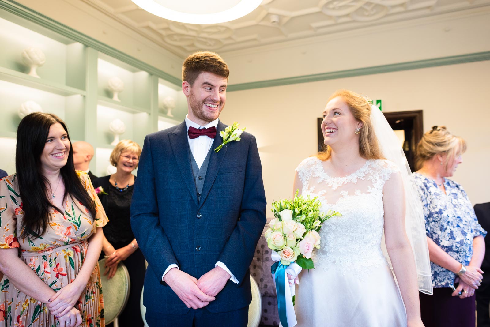 Belinda and Chris smile at each other at the bottom of the aisle in the Evelyn Room, Lewes Register Ofiice before their wedding.