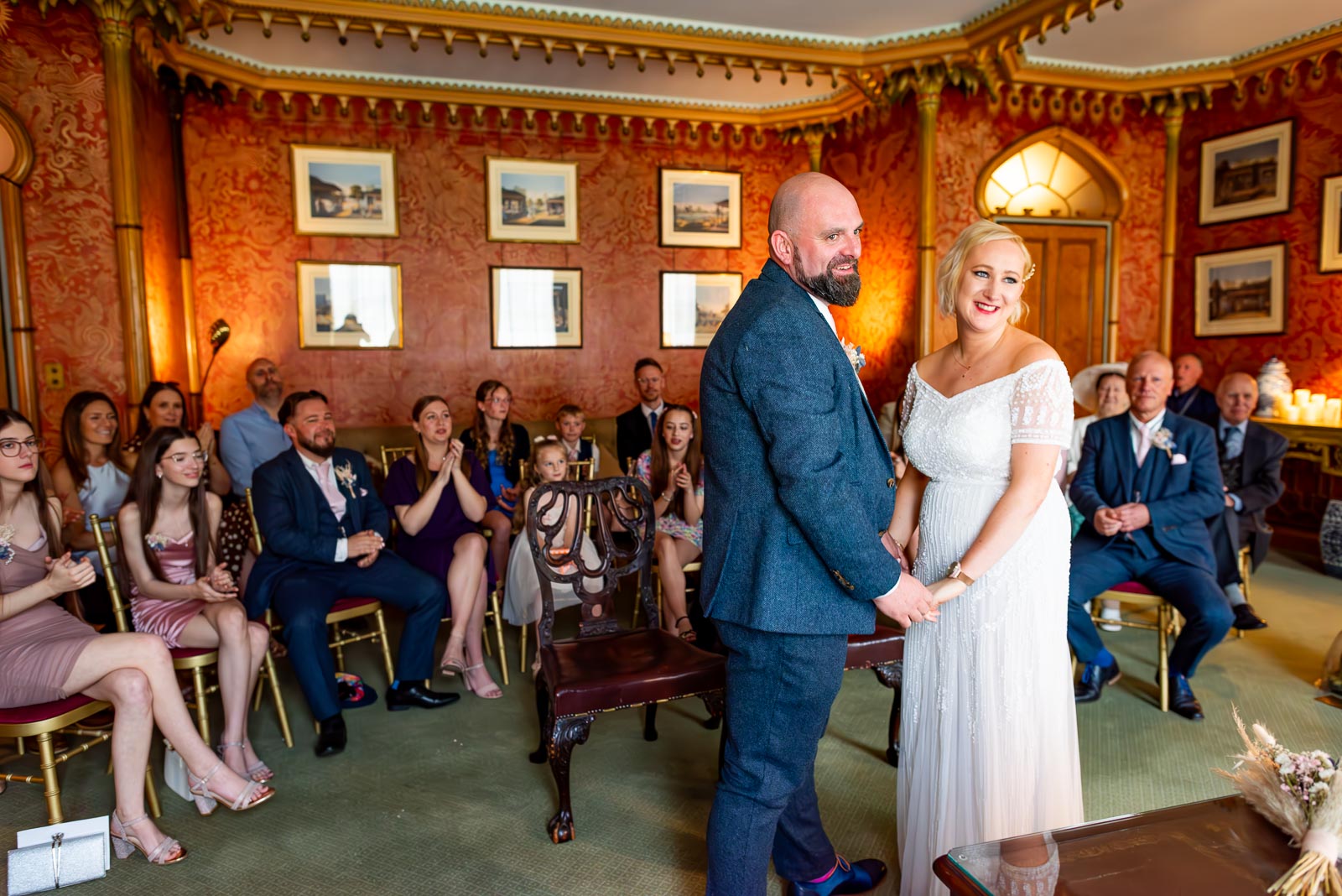 Georgina and Andy smile on front of their guests at The Red Drawing Room in Brighton Royal Pavilion after their wedding.